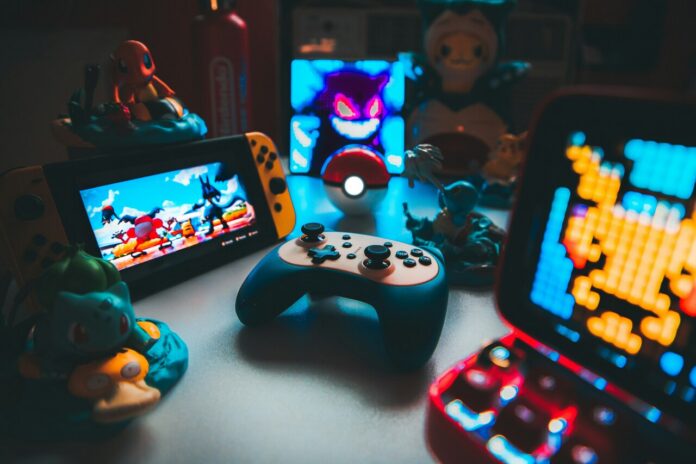 Mesa gamer com iluminação azul e vermelha reunindo um controle sem fio, console Nintendo Switch, miniaturas colecionáveis de Pokémon e uma Pokébola ao centro.